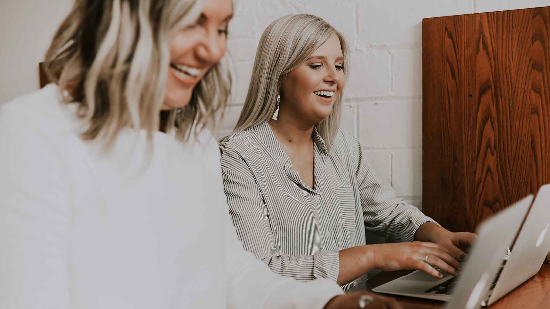 to lyshårede kvinder sidder og smiler mens de arbejder mod success